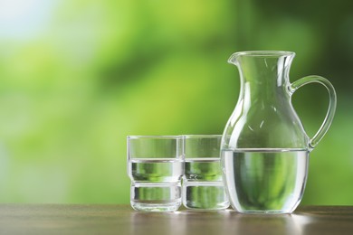 Photo of Jug and glasses with clear water on wooden table against blurred green background. Space for text