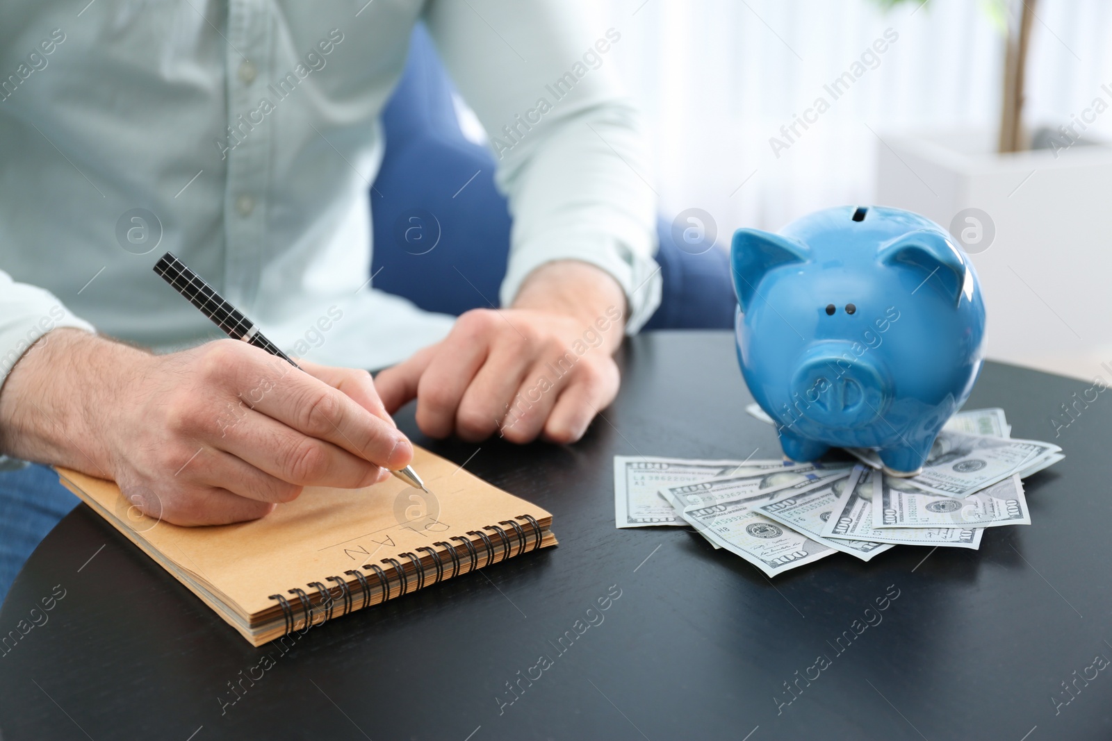 Photo of Businessman with notebook, piggy bank and money at table indoors, closeup