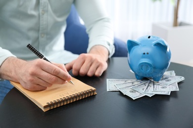 Businessman with notebook, piggy bank and money at table indoors, closeup