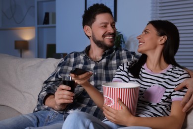 Photo of Happy couple spending time together at home in evening. Woman holding popcorn and changing TV channels with remote control