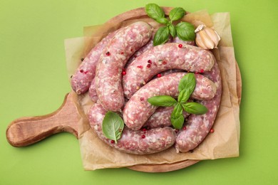 Photo of Board with raw homemade sausages, basil leaves and peppercorns on green table, top view