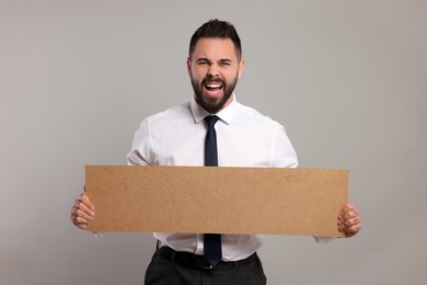 Photo of Angry young man holding blank cardboard banner on light grey background, space for text