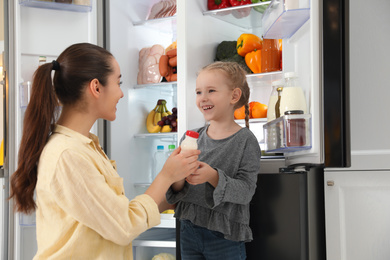 Photo of Young mother and her daughter with bottle of yoghurt near open refrigerator in kitchen
