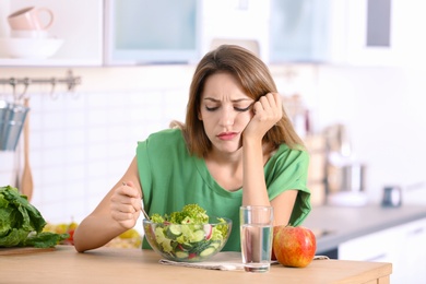 Photo of Unhappy woman eating vegetable salad at table in kitchen. Healthy diet