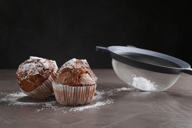 Sieve with sugar powder and muffins on grey textured table, closeup