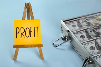 Economic profit. Easel with note and banknotes in briefcase on light blue background, closeup