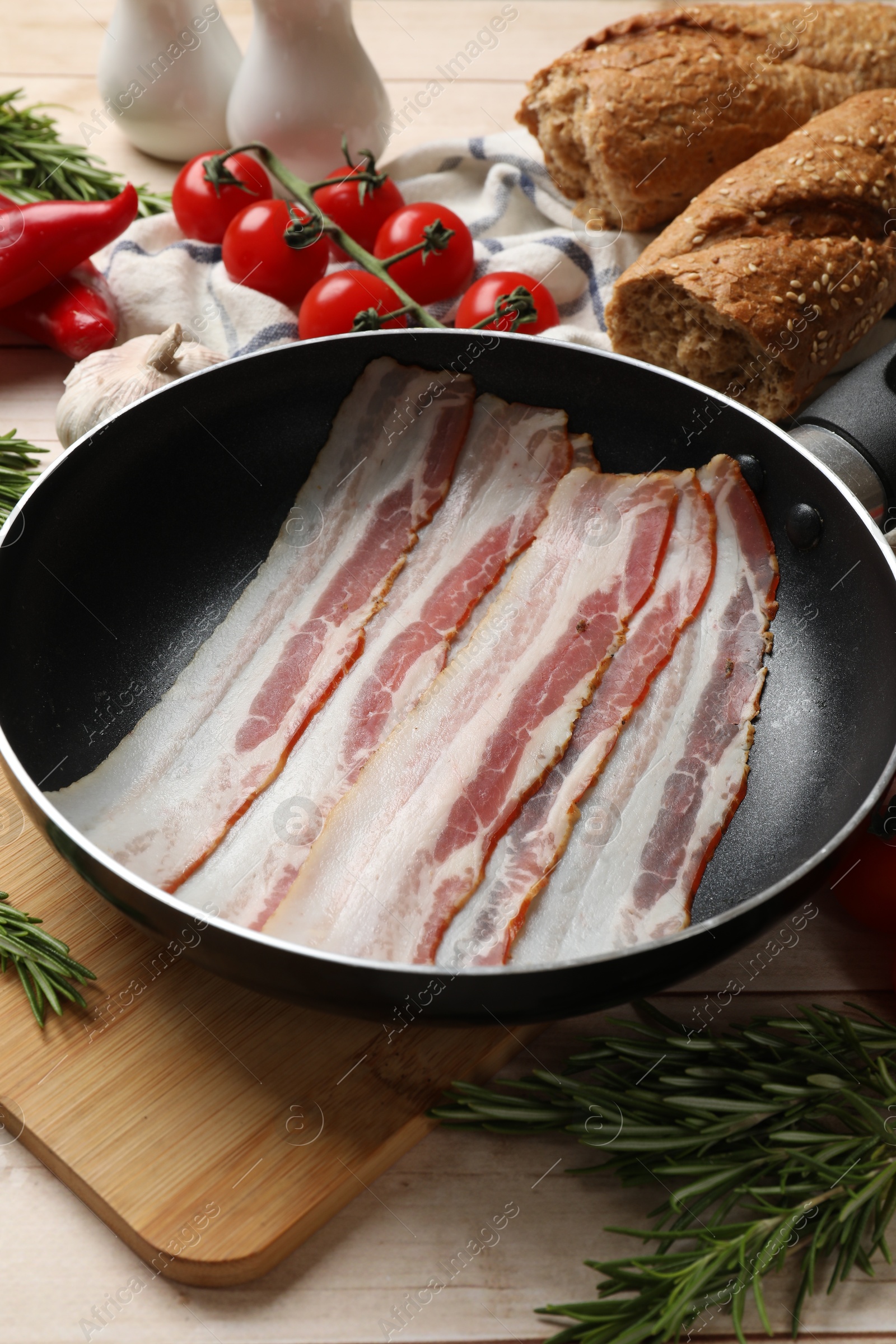 Photo of Slices of raw bacon in frying pan and fresh products on wooden table, closeup