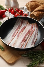 Slices of raw bacon in frying pan and fresh products on wooden table, closeup