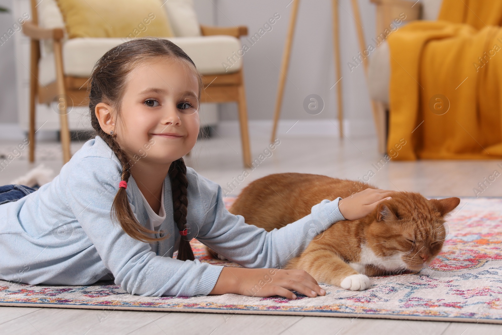 Photo of Smiling little girl petting cute ginger cat on carpet at home