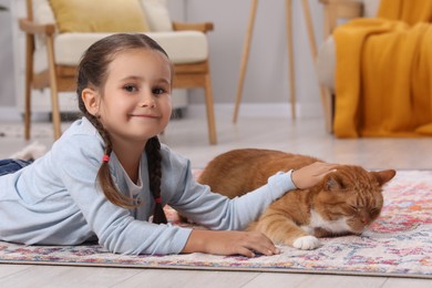 Smiling little girl petting cute ginger cat on carpet at home