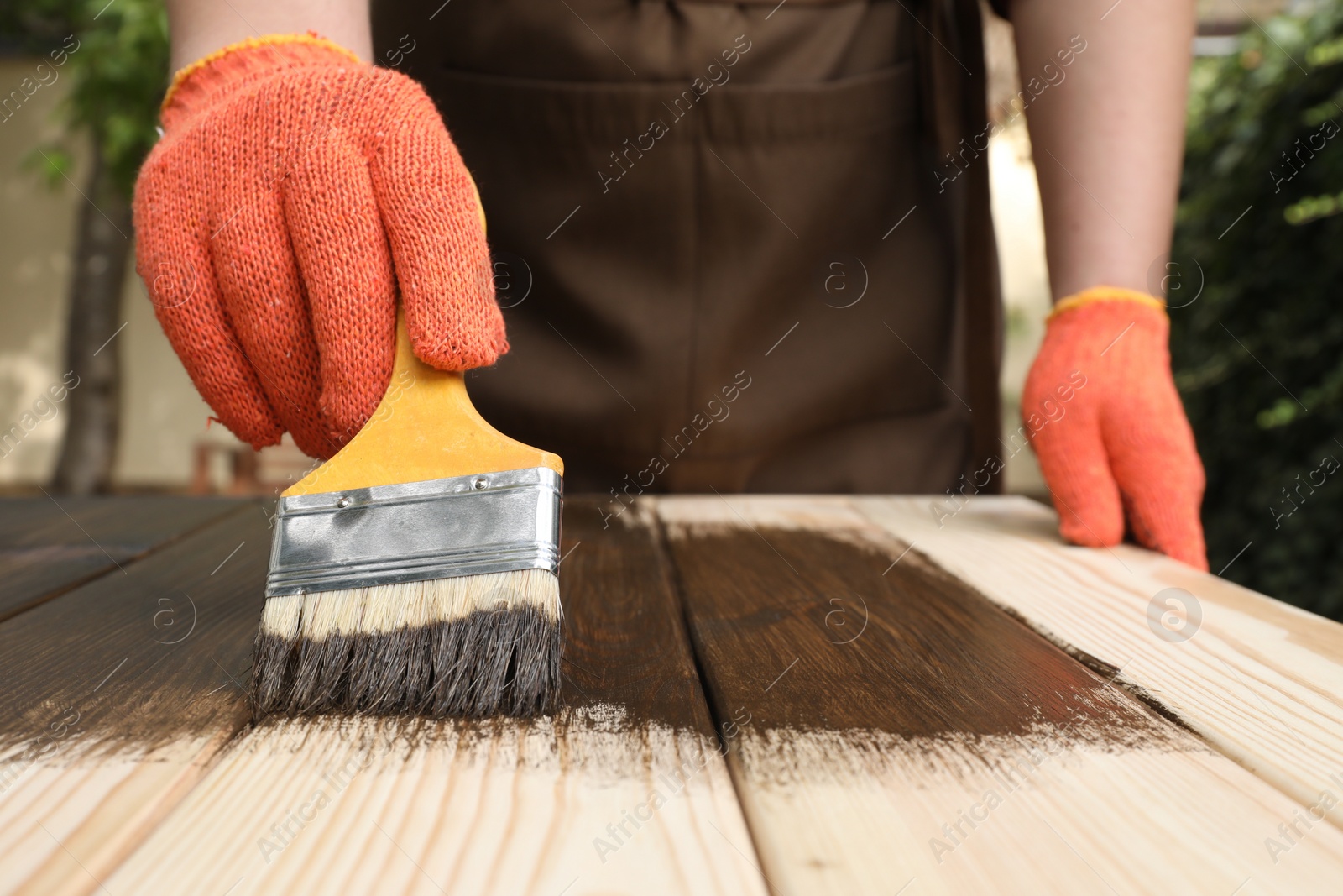 Photo of Man applying wood stain onto wooden surface against blurred background, closeup