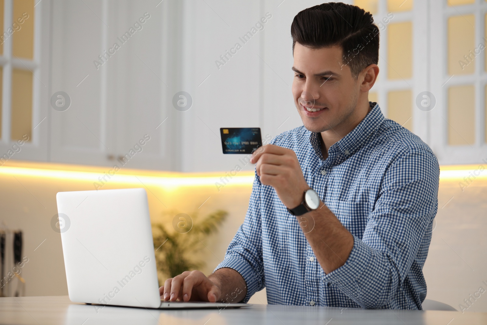 Photo of Man using laptop and credit card for online payment at table in kitchen