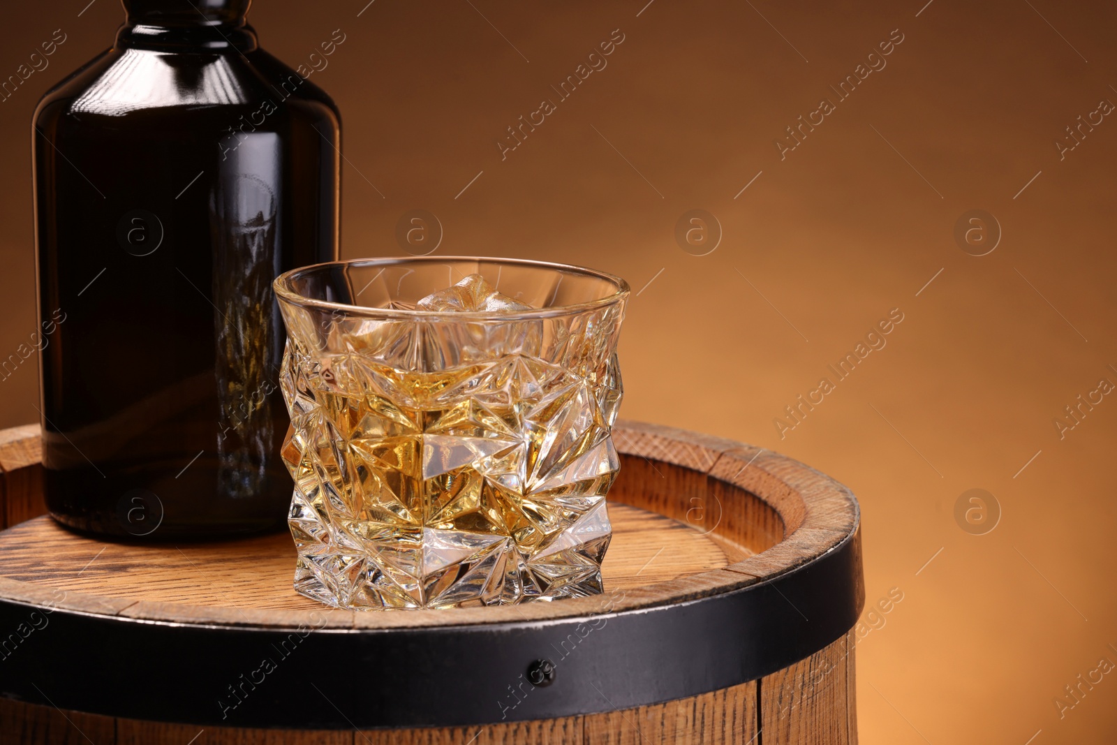 Photo of Whiskey with ice cubes in glass and bottle on wooden barrel against brown background, space for text