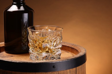 Whiskey with ice cubes in glass and bottle on wooden barrel against brown background, space for text