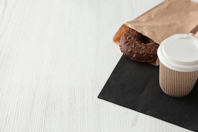 Tasty donut in paper bag and coffee on wooden table. Space for text