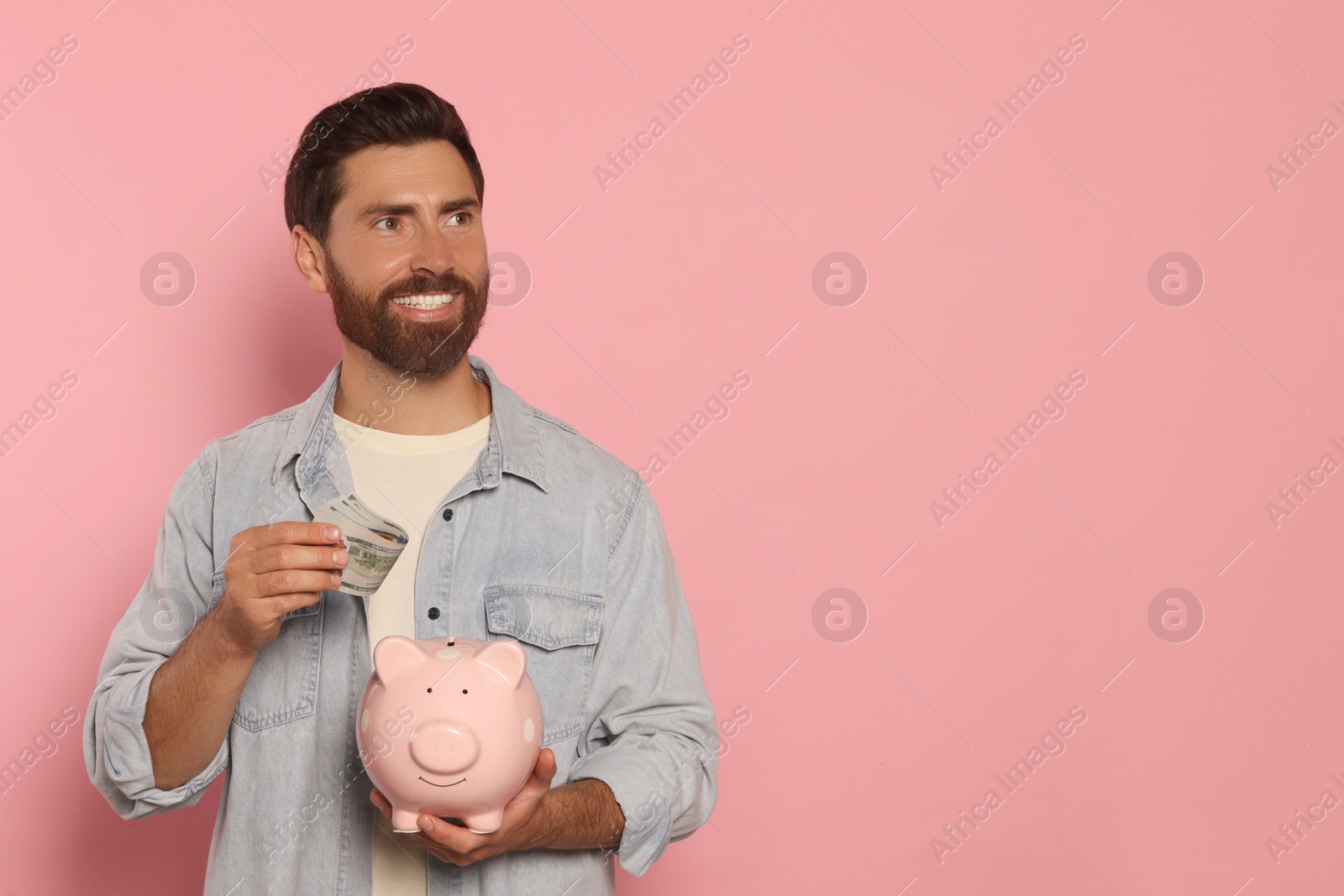 Photo of Happy man putting money into piggy bank on pale pink background, space for text