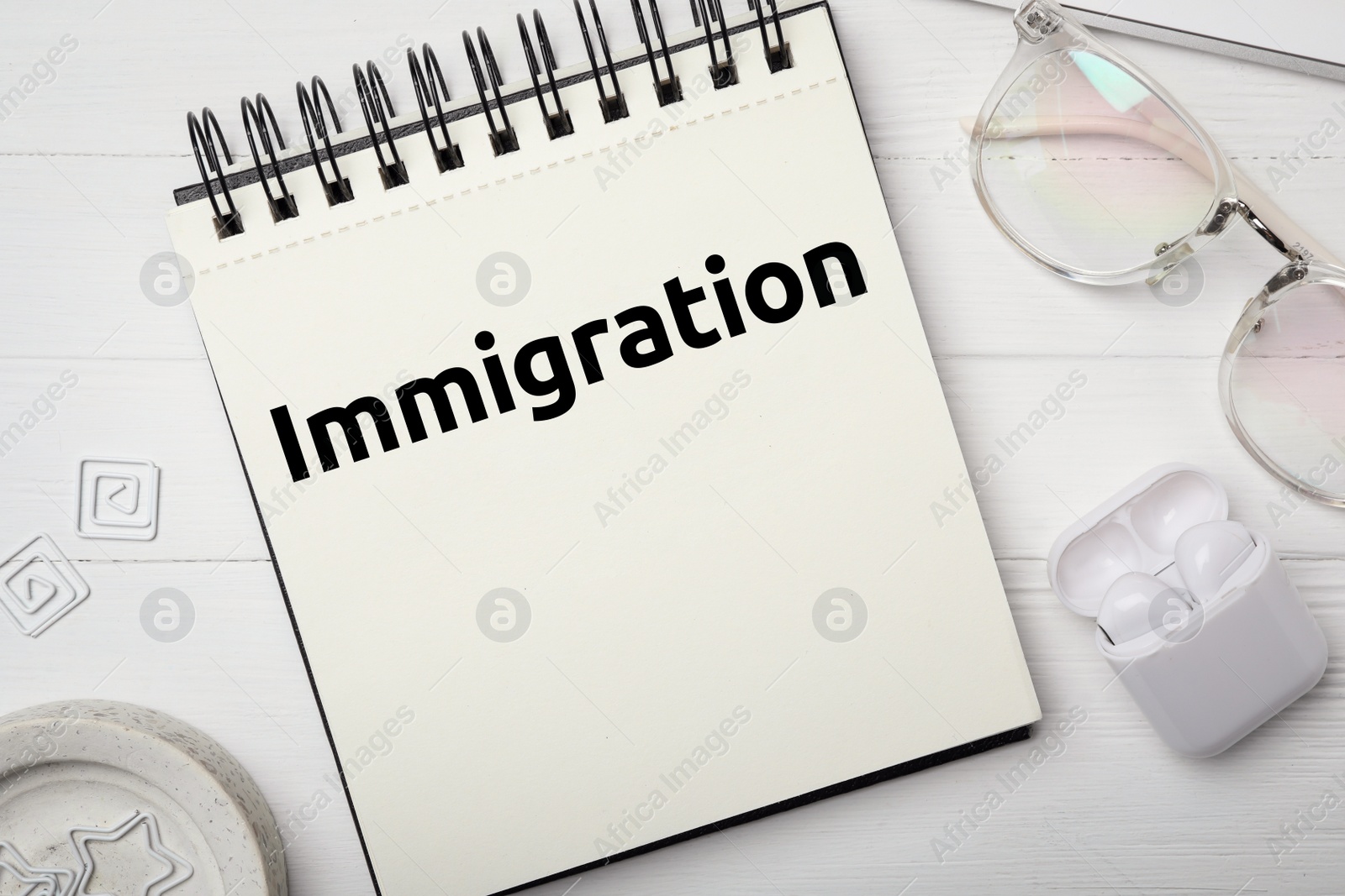 Image of Notebook with word IMMIGRATION, eyeglasses and earphones on white wooden table, flat lay