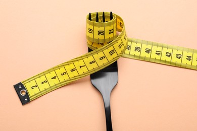 Fork with measuring tape on pale orange background, top view. Diet concept