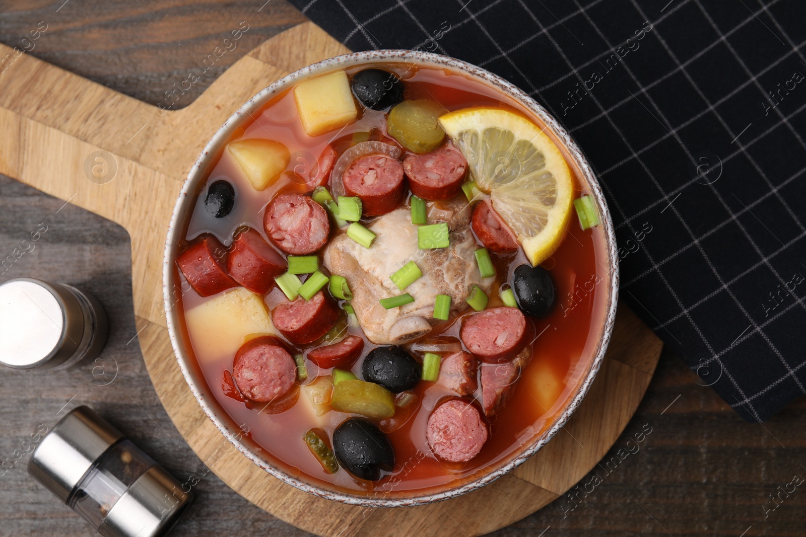 Photo of Meat solyanka soup with thin dry smoked sausages in bowl on wooden table, flat lay