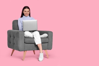 Happy woman with laptop sitting in armchair on pink background