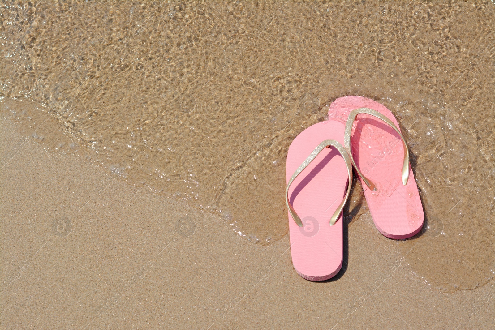 Photo of Stylish pink flip flops on wet sand getting hit by sea wave, above view. Space for text