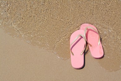 Photo of Stylish pink flip flops on wet sand getting hit by sea wave, above view. Space for text