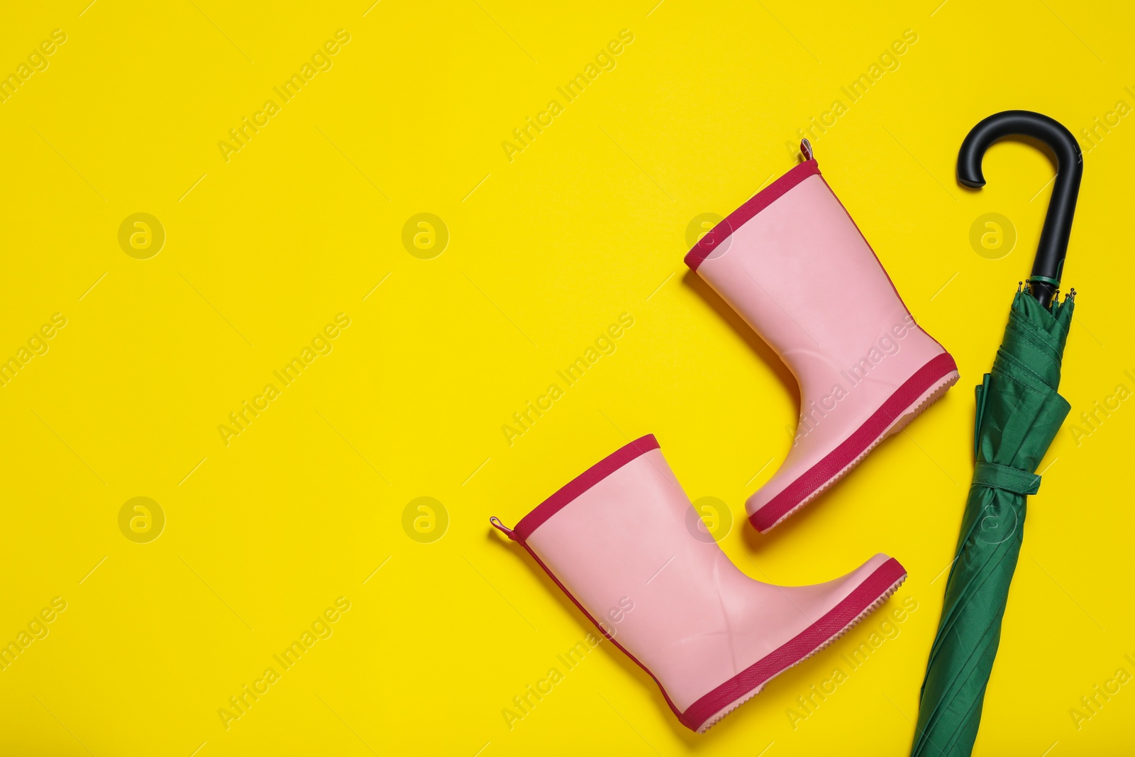 Photo of Pair of pink rubber boots near green umbrella on yellow background, flat lay. Space for text