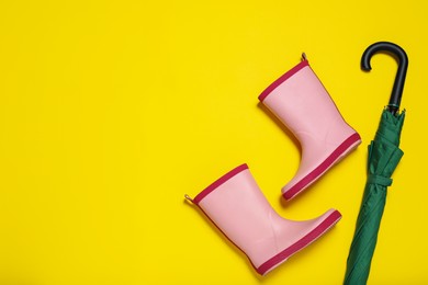 Photo of Pair of pink rubber boots near green umbrella on yellow background, flat lay. Space for text
