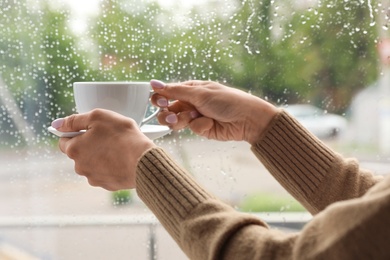 Young woman with cup of coffee near window indoors on rainy day, closeup