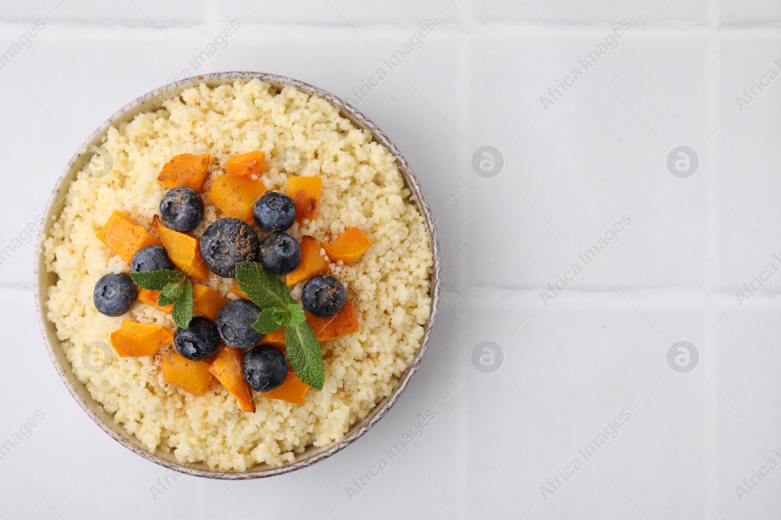 Photo of Bowl of tasty couscous with blueberries, pumpkin and mint on white tiled table, top view. Space for text