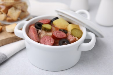 Meat solyanka soup with thin dry smoked sausages in pot on light table, closeup