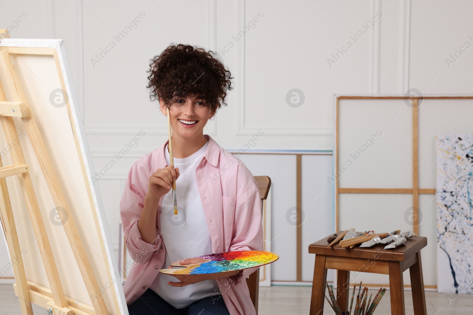 Photo of Young woman holding brush and artist`s palette near easel with canvas