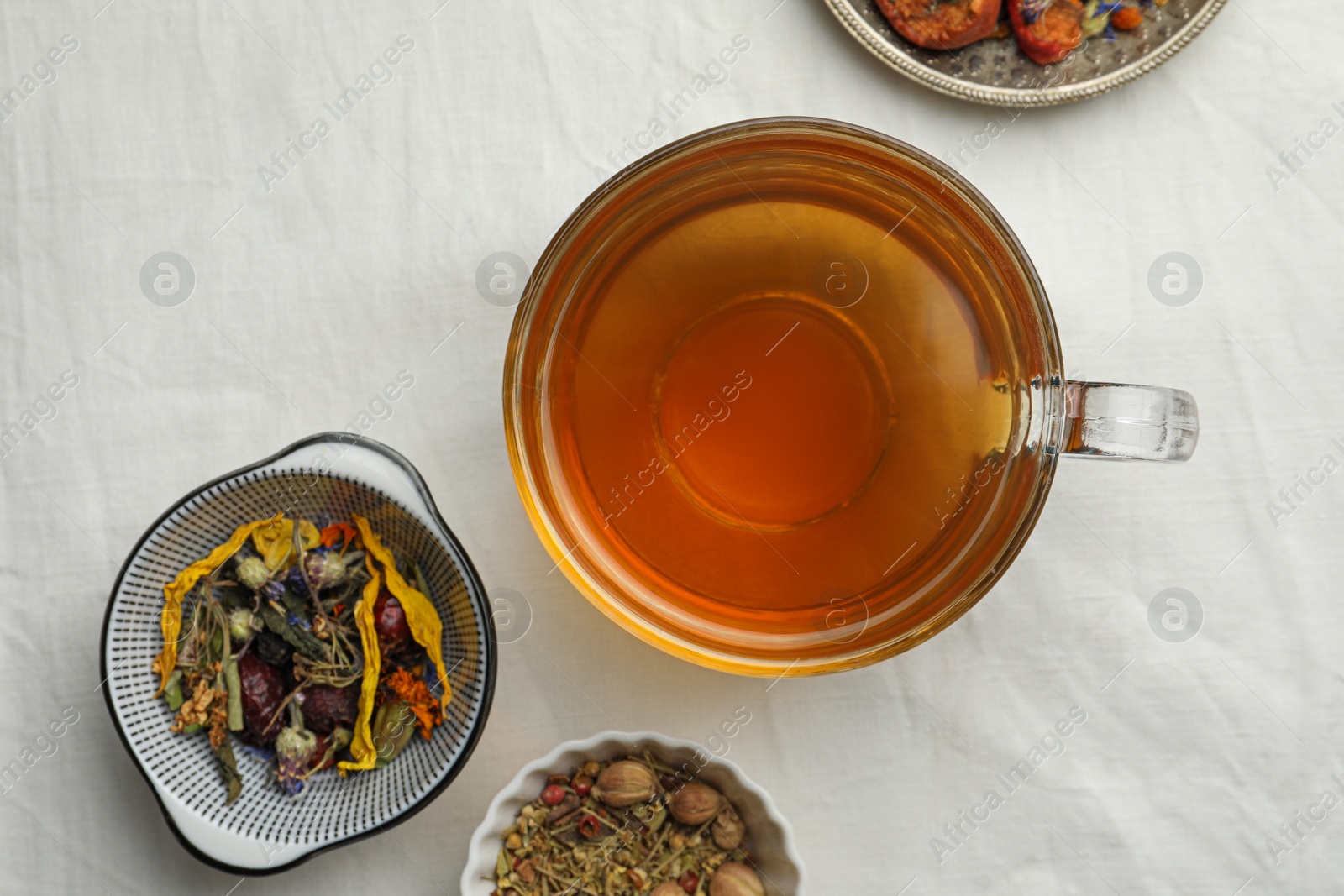 Photo of Cup of freshly brewed tea, dried herbs and berries on white fabric, flat lay