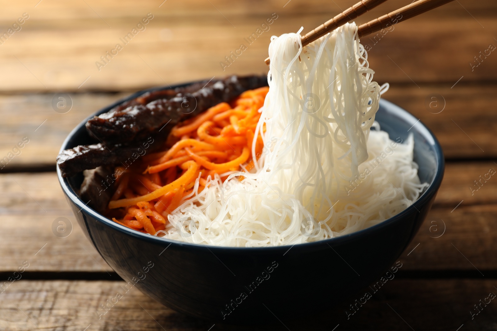 Photo of Chopsticks with tasty cooked rice noodles over bowl on wooden table, closeup