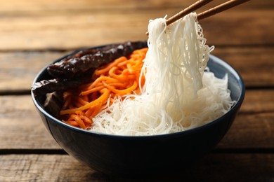 Photo of Chopsticks with tasty cooked rice noodles over bowl on wooden table, closeup