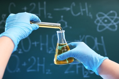 Photo of Scientist pouring liquid into flask against chalkboard, closeup. Chemistry glassware
