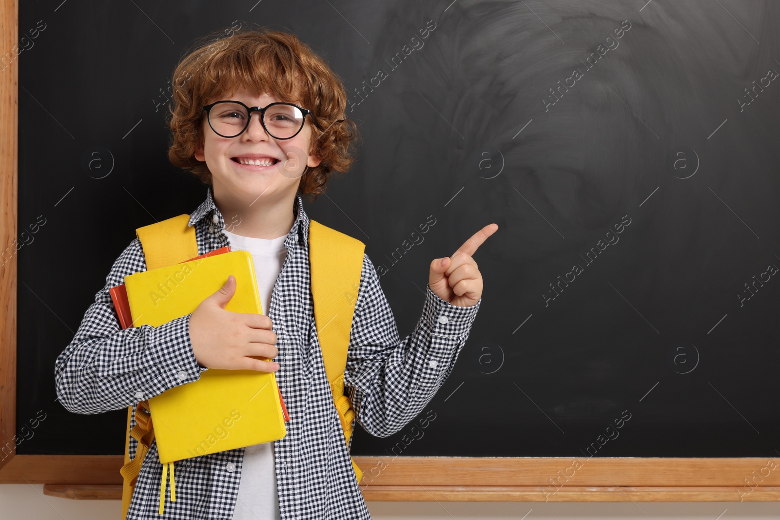 Photo of Happy schoolboy in glasses with backpack and books pointing at something near blackboard, space for text
