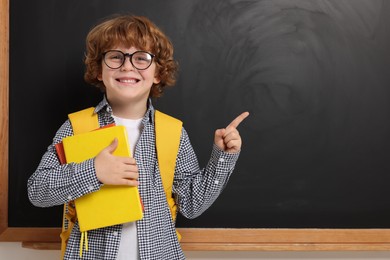 Happy schoolboy in glasses with backpack and books pointing at something near blackboard, space for text