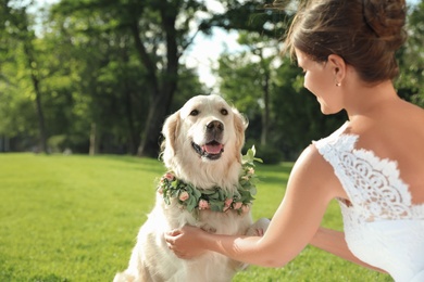 Photo of Bride and adorable Golden Retriever wearing wreath made of beautiful flowers on green grass outdoors