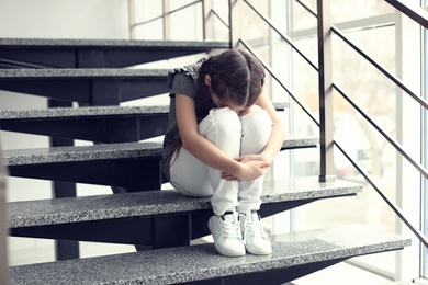 Photo of Depressed little girl sitting on stairs indoors. Time to visit child psychologist