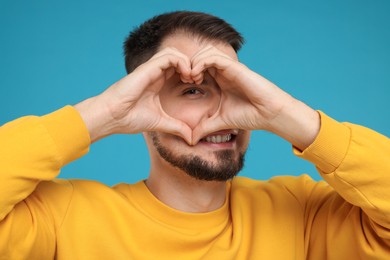 Man looking through folded in shape of heart hands on light blue background