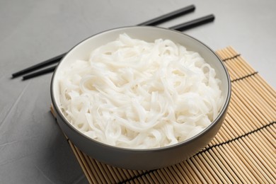 Bowl with cooked rice noodles, straw mat and chopsticks on light grey table, closeup