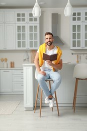 Photo of Handsome man with book sitting on stool in kitchen