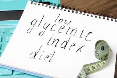 Photo of Notebook with words Low Glycemic Index Diet, measuring tape and calculator on table, closeup