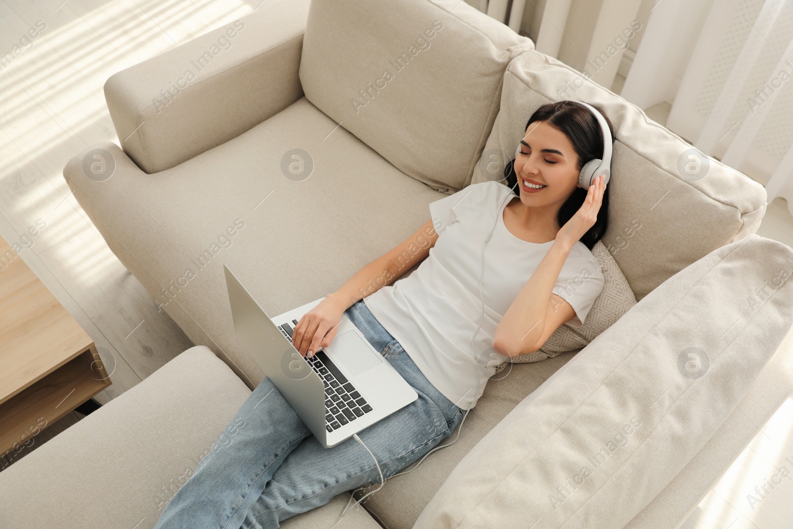 Photo of Woman with laptop and headphones lying on sofa at home