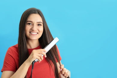 Young woman using hair iron on blue background, space for text