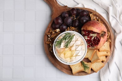 Photo of Board with tasty baked camembert, croutons, grapes, walnuts and pomegranate on white tiled table, top view. Space for text