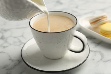 Pouring milk from pitcher into cup with coffee at white marble table, closeup