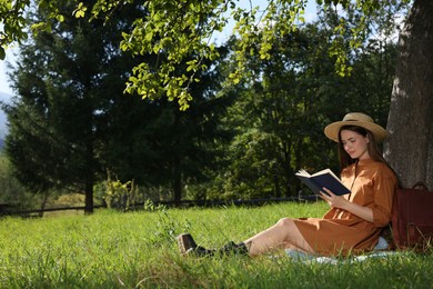 Photo of Young woman reading book under tree on meadow near forest