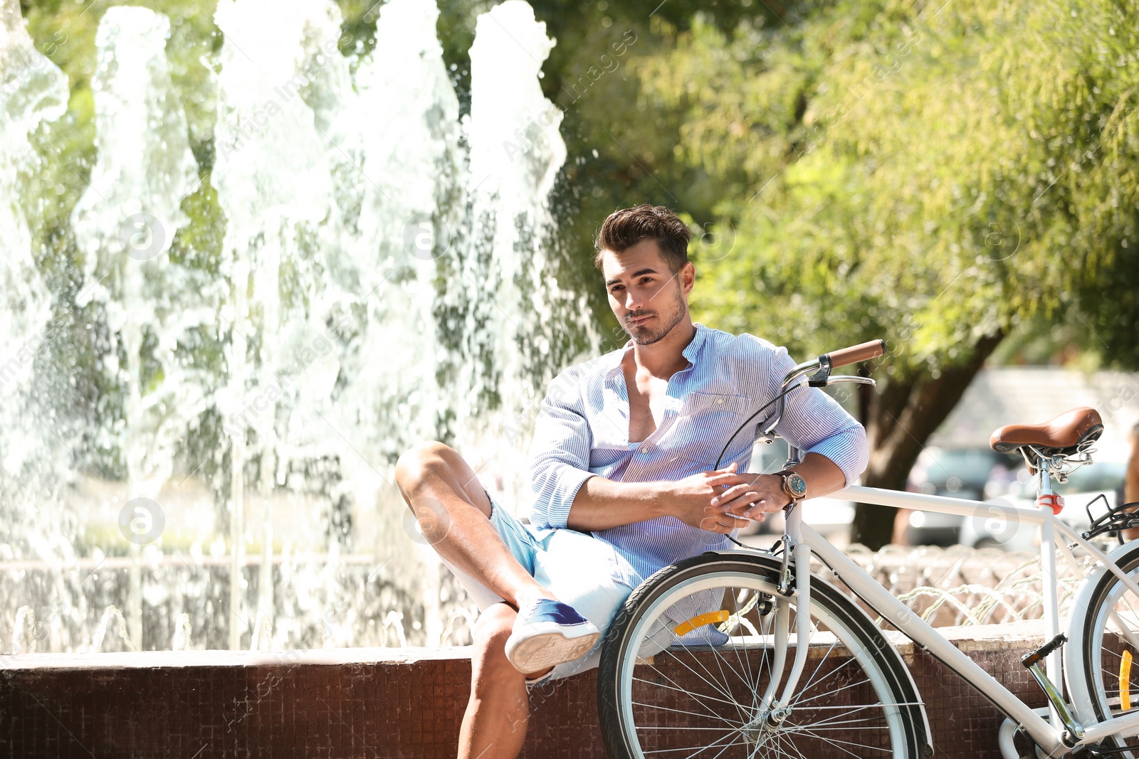 Photo of Handsome young hipster man sitting with bicycle near fountain in park